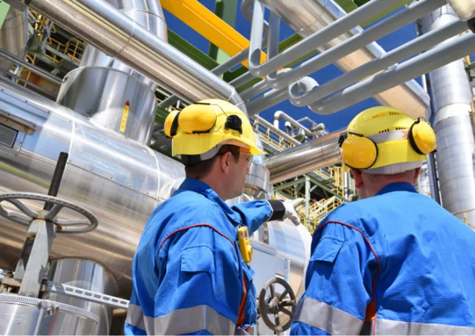 Two men in blue overalls and yellow safety helmets working on a large industrial pipe